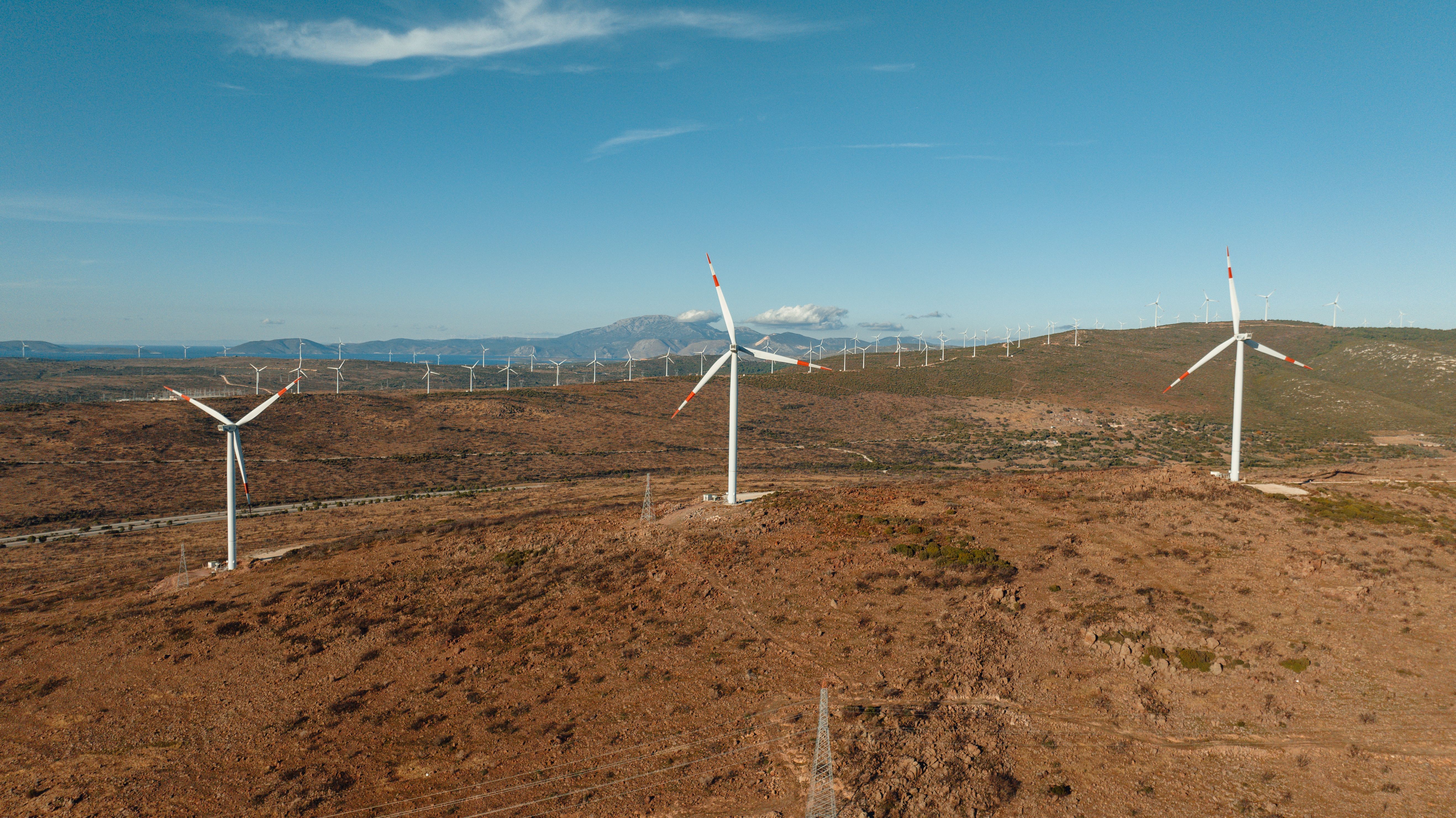 Oblique aerial view over dry, scrubby hills with a line of wind turbines and mountains on the horizon.